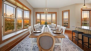 Een eetkamer met een lange tafel gedekt voor zes personen, grote ramen en uitzicht op bomen.
