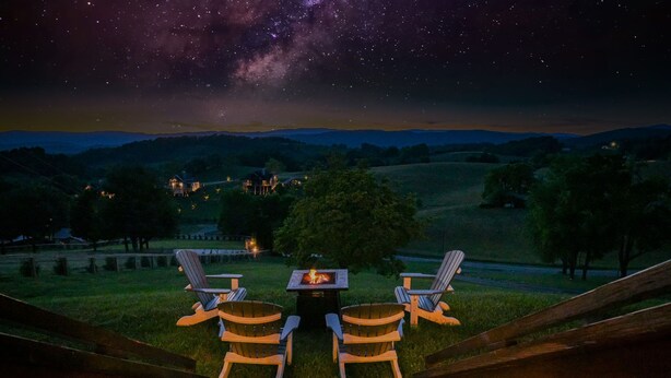 Een nachthemel met sterren en de Melkweg boven een begroeiend landschap met twee Adirondack-stoelen en een vuurkuil.