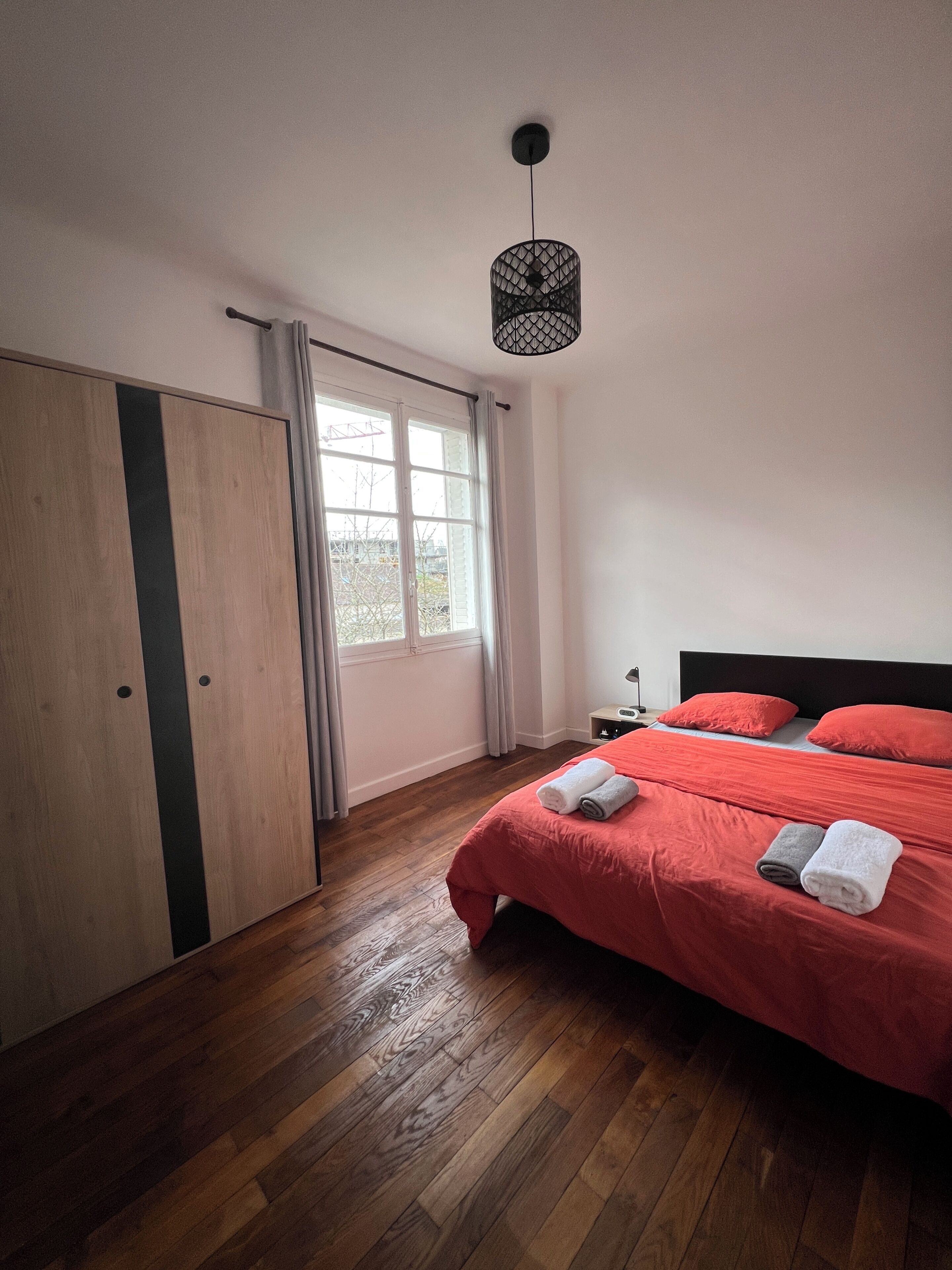 Un dormitorio con un armario de madera, una cama con ropa de cama roja, una lámpara colgante y una ventana con cortinas.