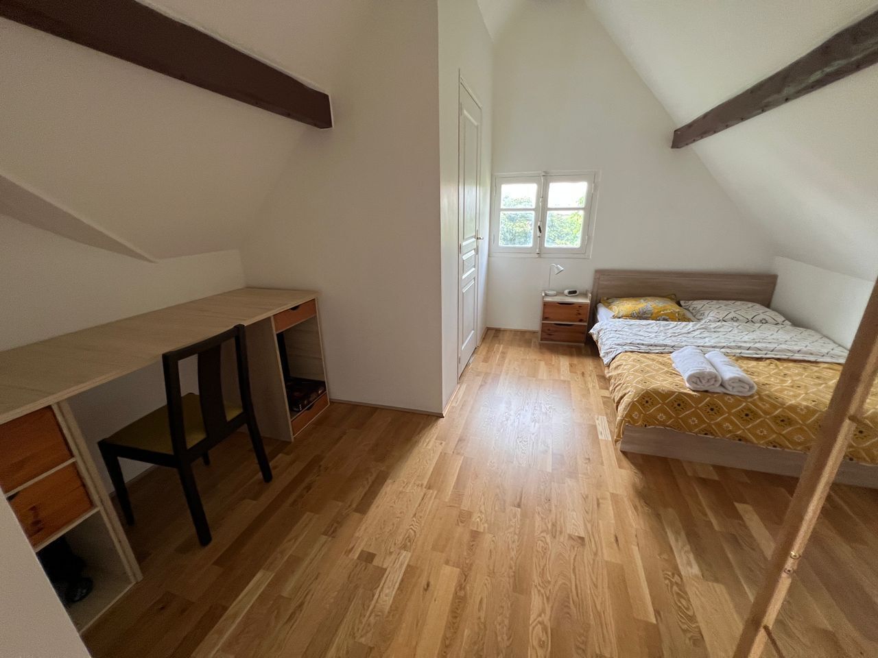 A bedroom with a wooden floor, a bed with a yellow patterned bedspread, a desk with a chair, and a window.