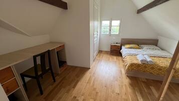 A bedroom with a wooden floor, a bed with a yellow patterned bedspread, a desk with a chair, and a window.