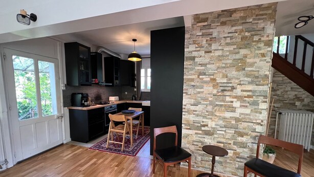 Uma sala ampla com piso de madeira, parede em pedra e vista para a cozinha.