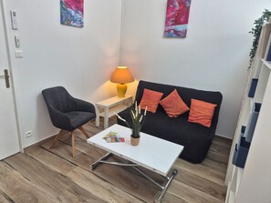 Una sala de estar pequeña y moderna, con un sofá negro, una mesa de centro blanca, un sillón negro, una mesita blanca y una lámpara amarilla.