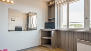 A room with a large mirror, a television on a shelf, a radiator, and a window with a view of buildings and trees. - Cozy Studio in the Heart of Avignon (Avignon)
