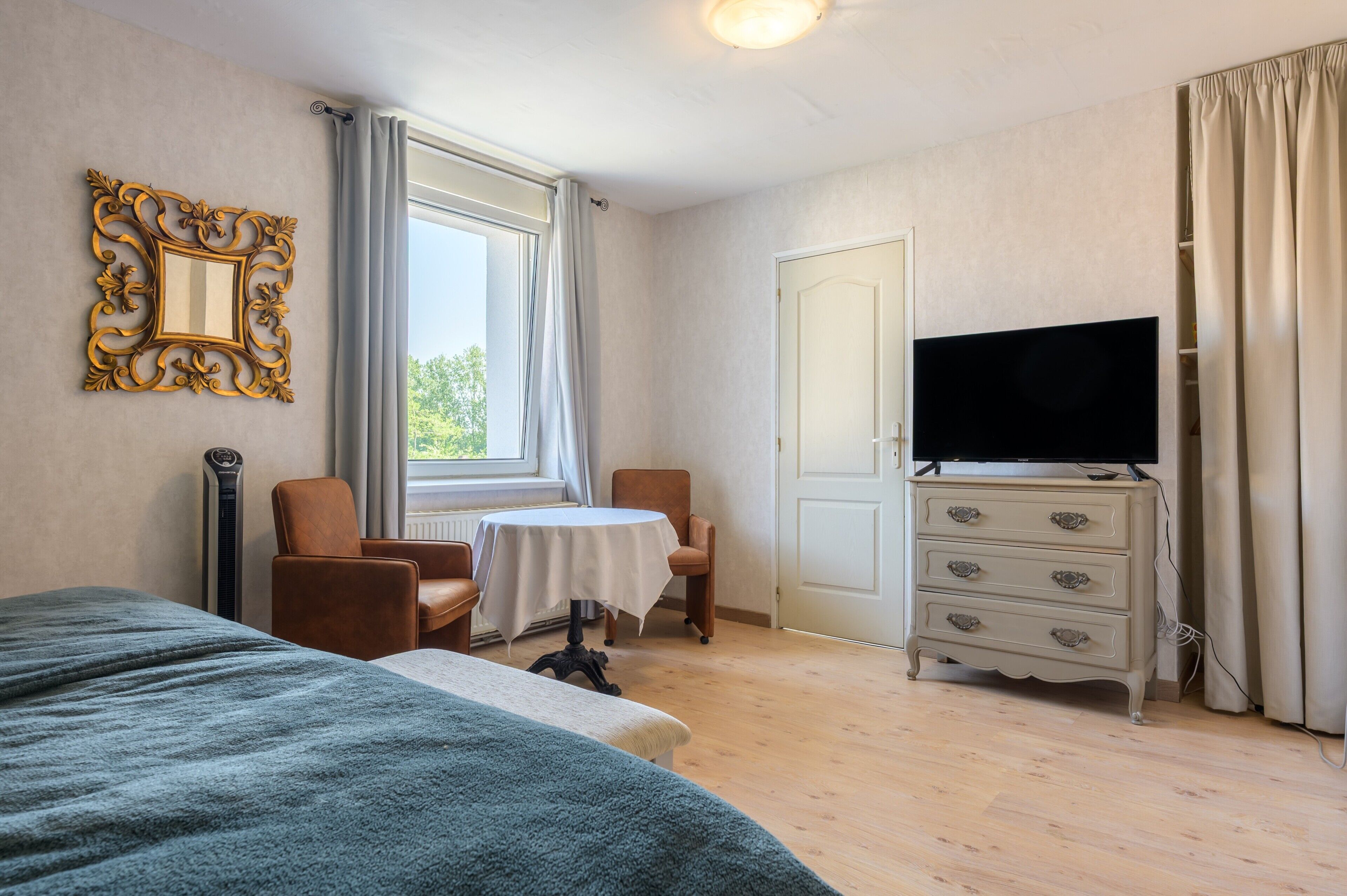 A hotel room with a bed, a television on a dresser, a small table with chairs, and a window with curtains.