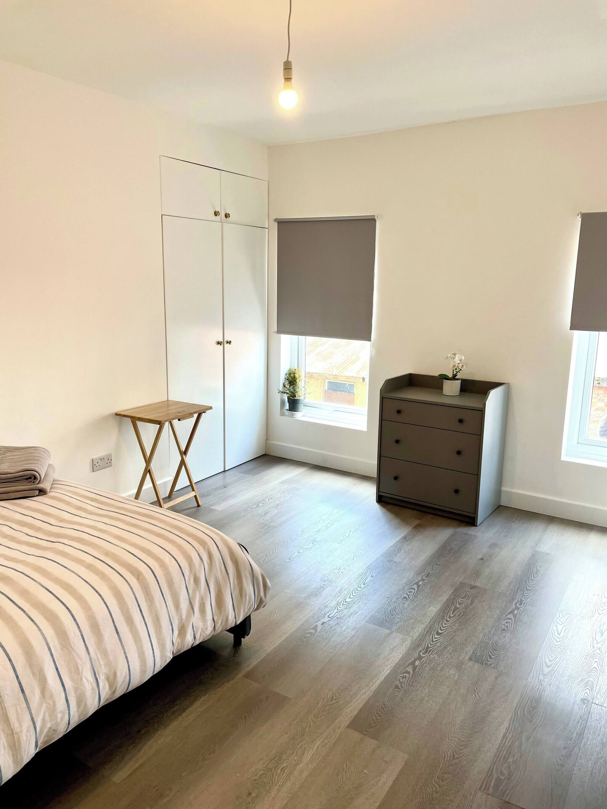 A bedroom with a bed, a dresser, a small table, a window with a blind, and a light fixture.