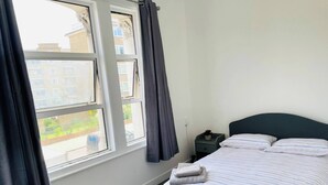 Quarto com cama, mesa de cabeceira, janela com cortinas e vista para prédios.