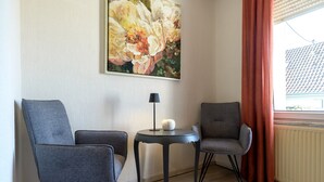Um quarto com duas cadeiras cinzentas, uma mesinha redonda com abajur, um quadro na parede e uma janela com cortinas.