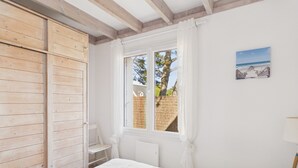 Un dormitorio con un armario de madera, una cama con edredón blanco, una ventana con vistas a un árbol y una valla, y un cuadro de una playa.