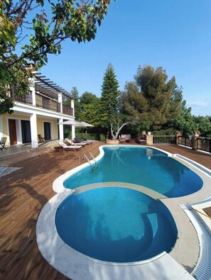 A swimming pool with a wooden deck, lounge chairs, and a building with a balcony. - Cave Luxury Villas Skiathos (Troulos)