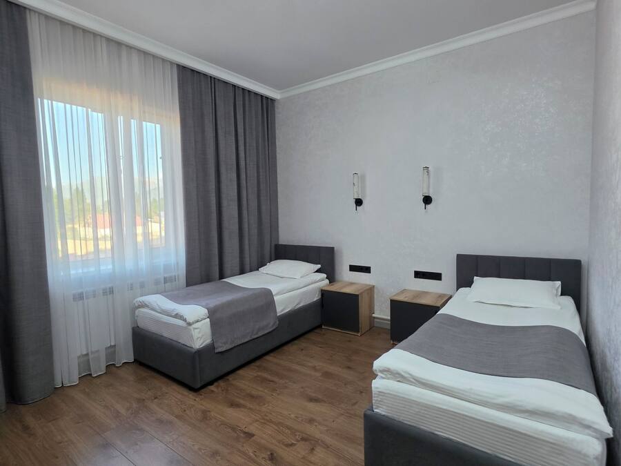 A modern hotel room with two beds, a wooden floor, a large window with curtains, and a wall-mounted light fixture.