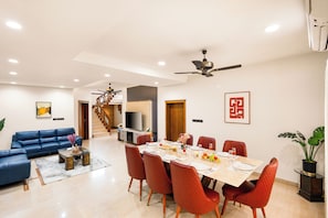 A spacious living area with a blue sofa, a dining table set for six, and a staircase leading to an upper floor. - ELIVAAS Valiyaveetil Mansion 5 BHK (Kanayannur)