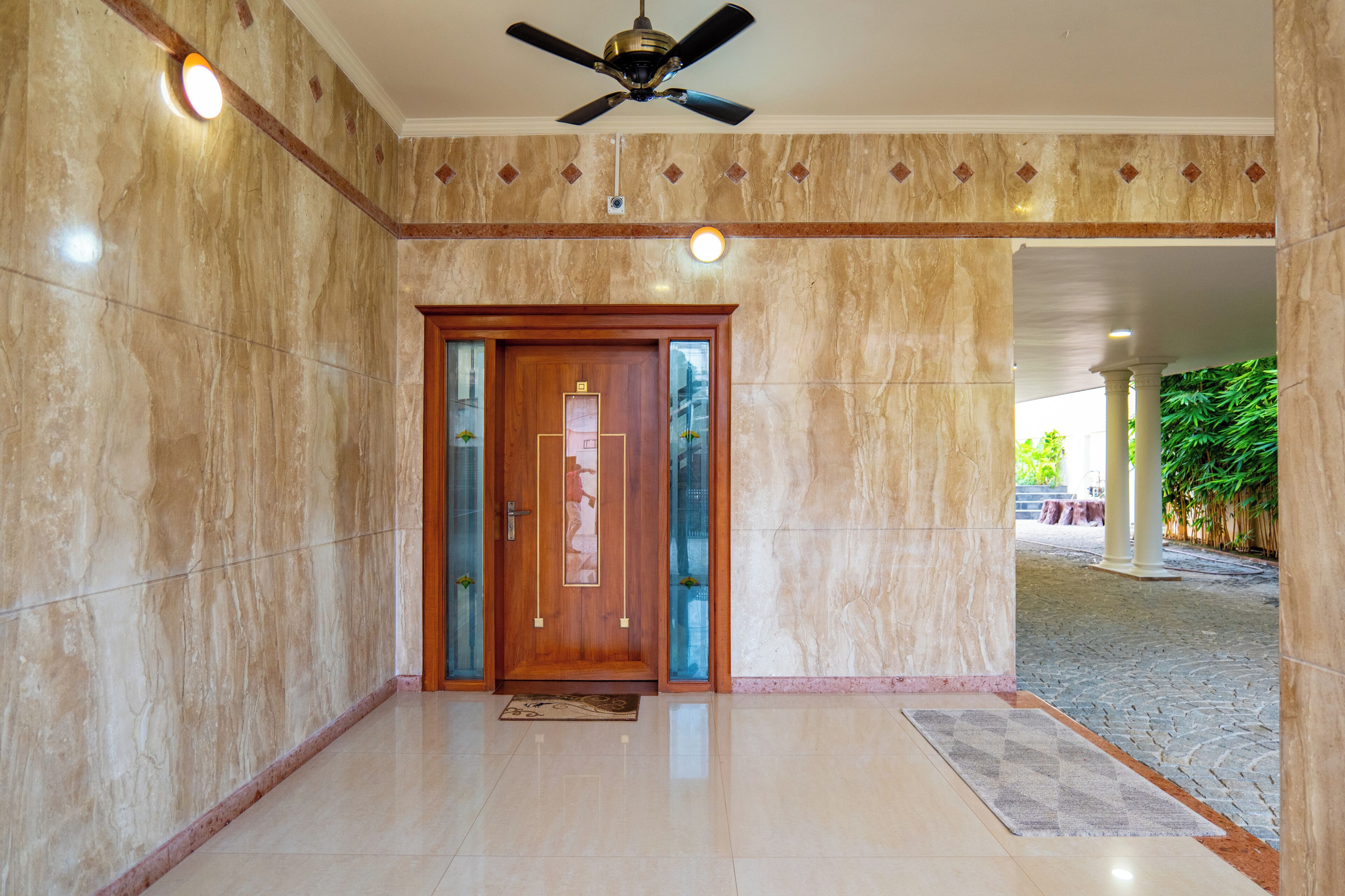 A lobby with a wooden door, marble walls, a ceiling fan, and a stone pathway leading outside.