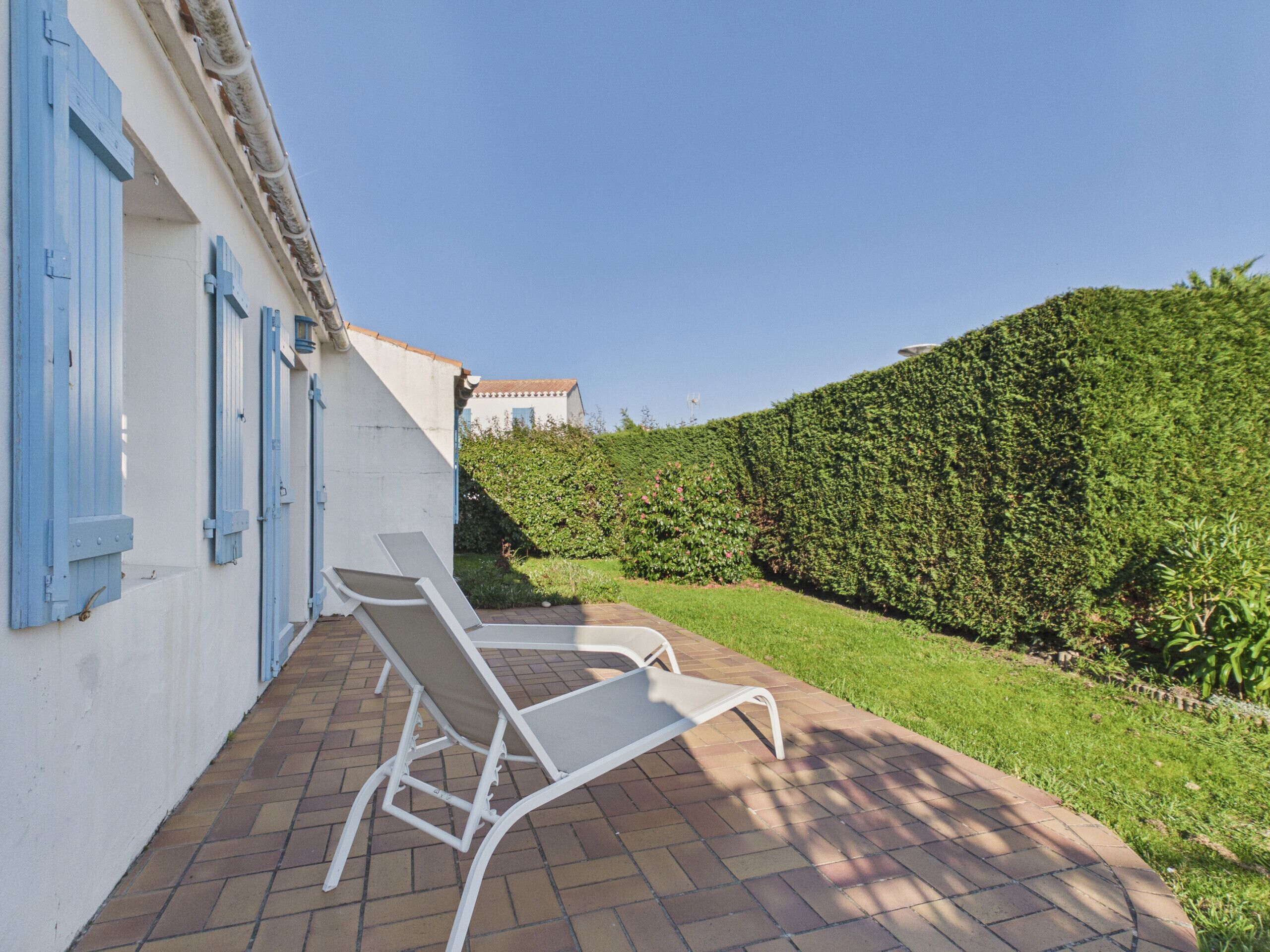 Eine Terrasse mit zwei weißen Liegestühlen, ein weißes Gebäude mit blauen Fensterläden und ein ordentlich geschnittener Heckenstreifen.