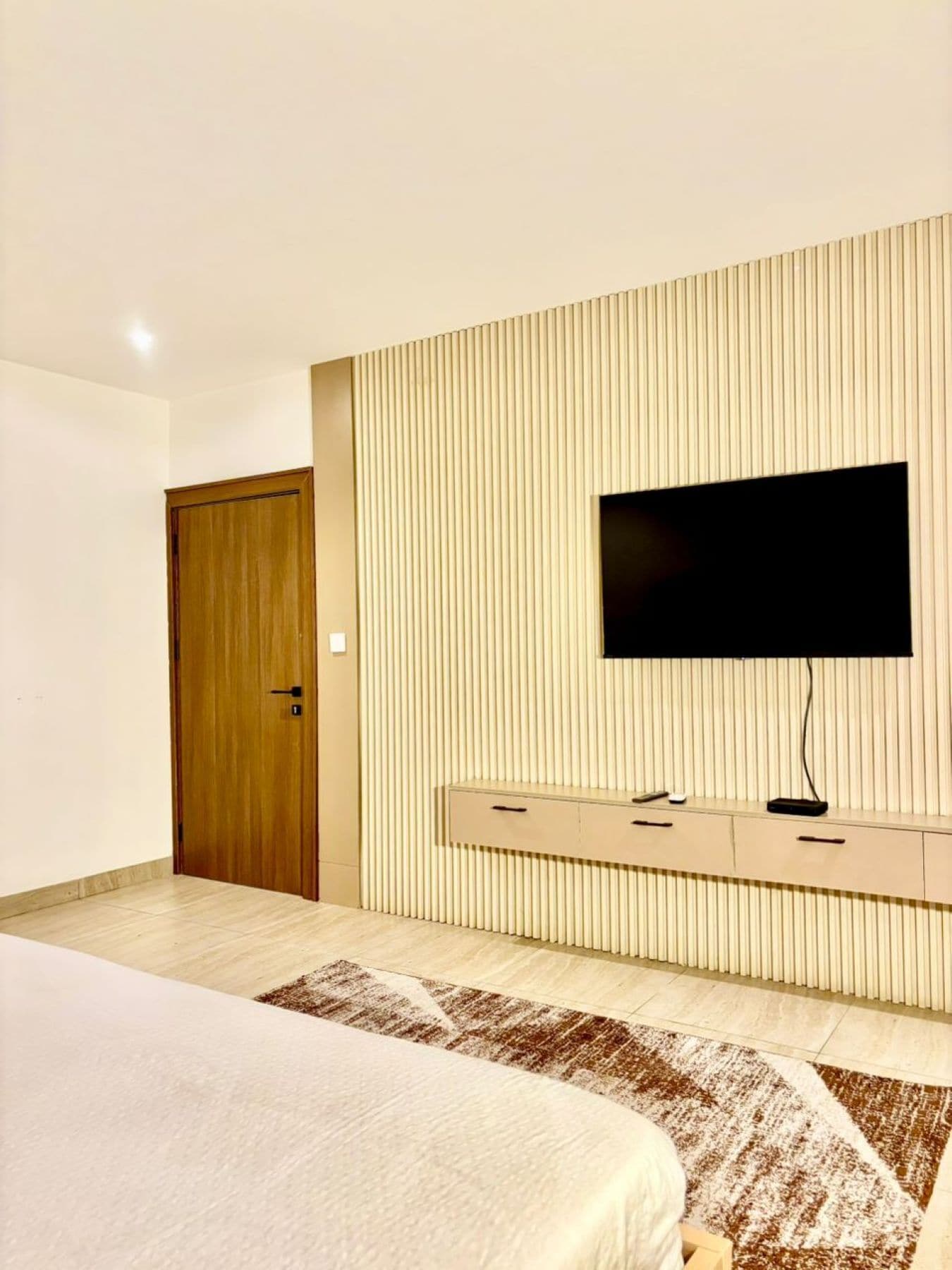 A bedroom with a bed, a TV mounted on a wall, a wooden door, and a patterned rug.