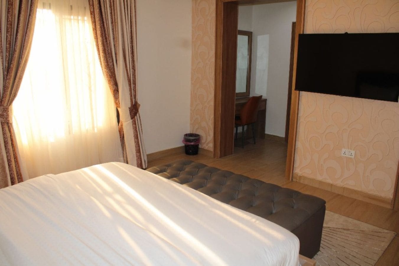 A bedroom with a bed, a small ottoman, a TV, and a dining area visible through an open door.