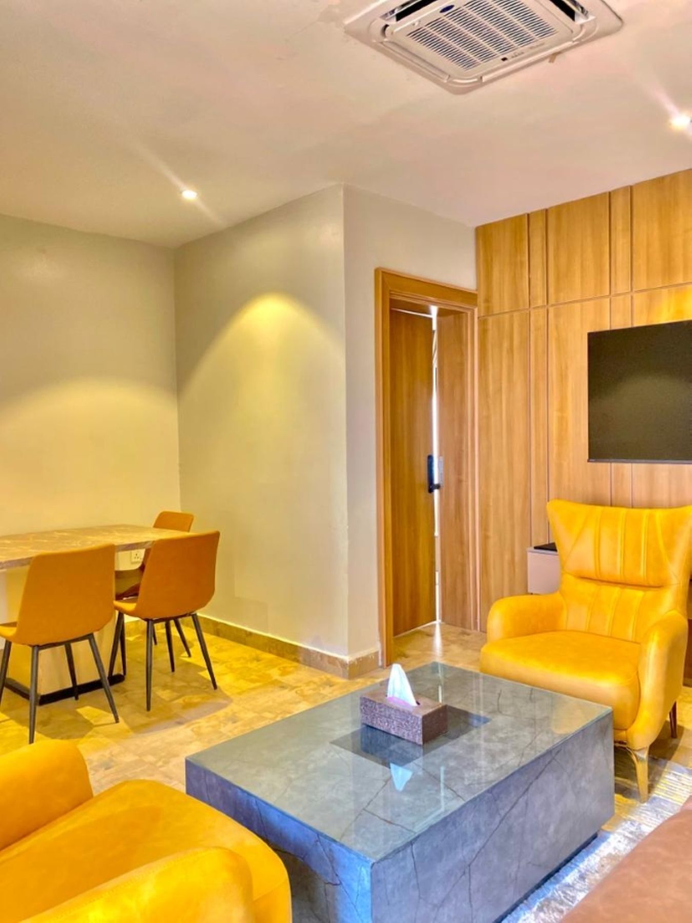 Una sala moderna con comedor, un televisor de pantalla plana en la pared y un sillón amarillo brillante.
