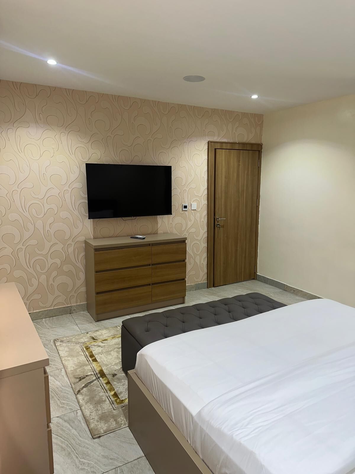 A bedroom with a bed, a TV mounted on the wall, a wooden wardrobe, and a patterned rug.
