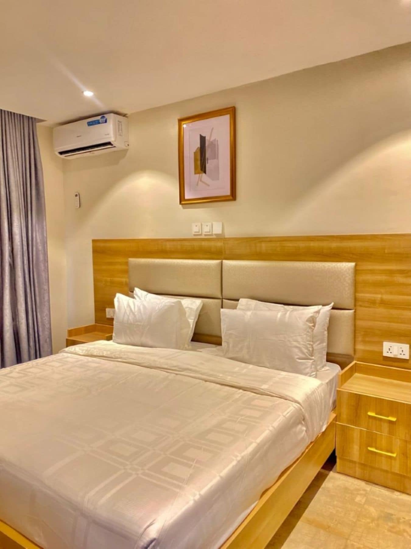 Quarto de hotel com uma cama grande, cabeceira de madeira, roupa de cama branca, ar condicionado, um quadro na parede e uma mesa de cabeceira com abajur.