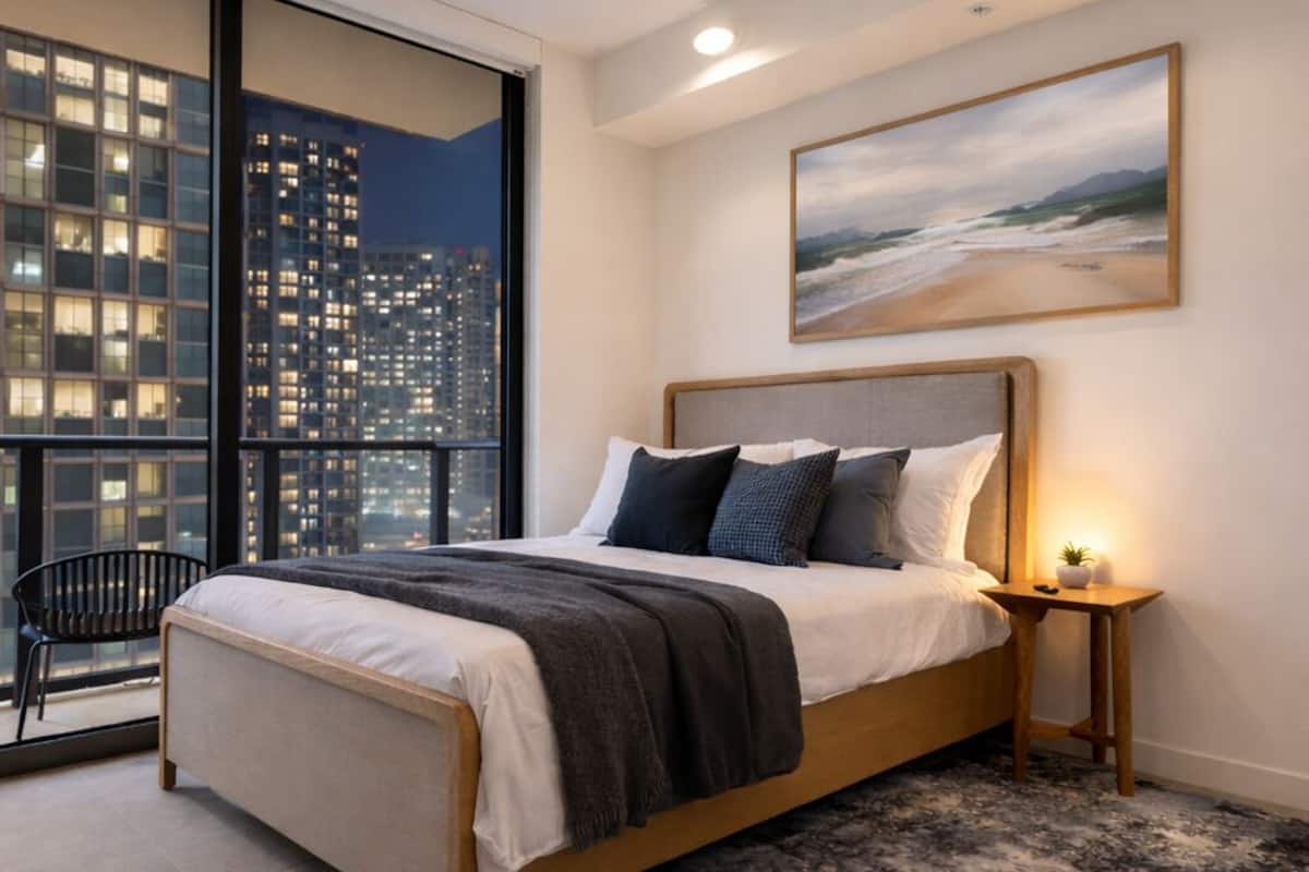 Un dormitorio con una cama grande, una mesita de noche, una silla, vistas a la ciudad a través de amplios ventanales y un cuadro en la pared.