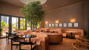 A modern lounge area with sofas, tables, and chairs, featuring a large indoor plant and framed pictures on the wall. - RS Suites - Miami World Center (Miami)