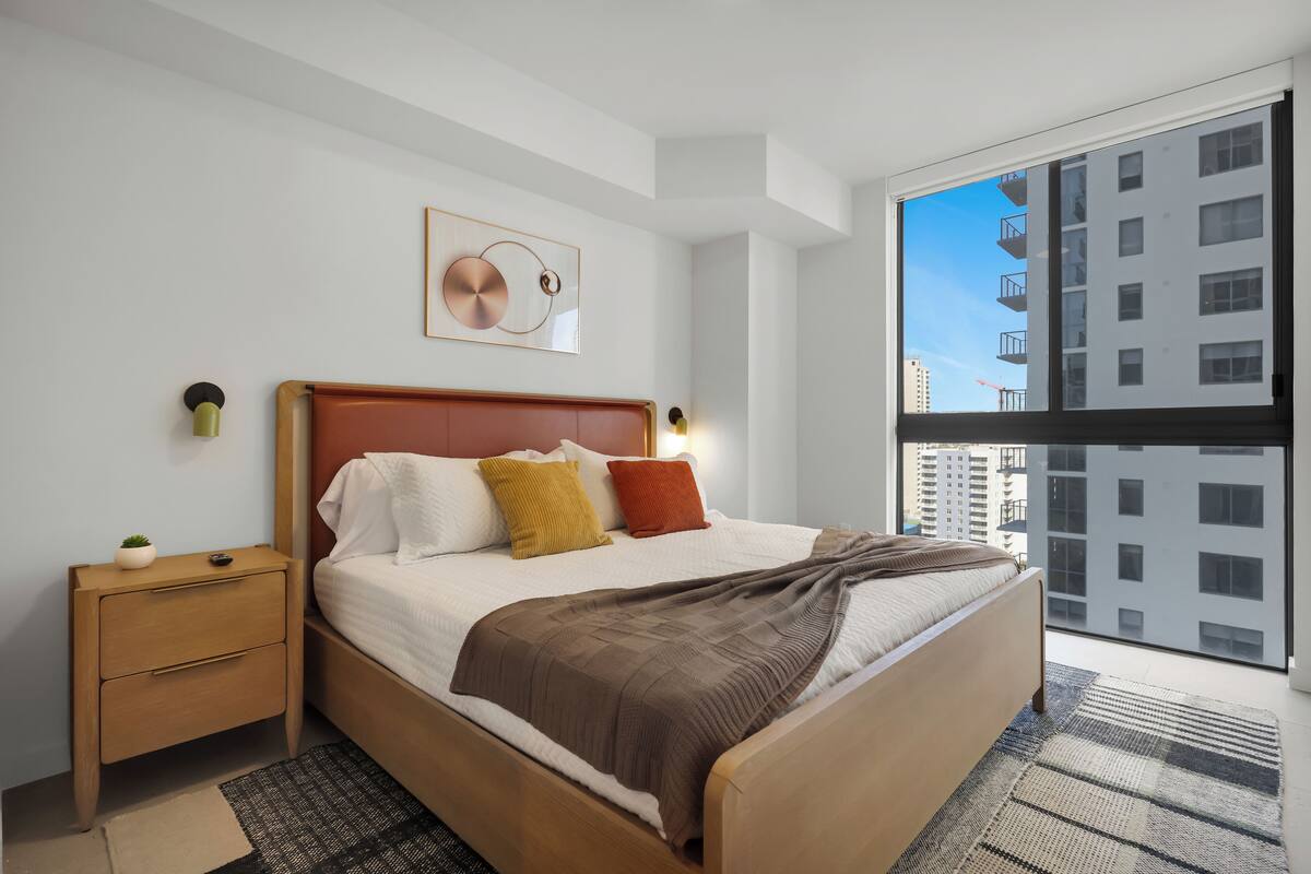 Un dormitorio moderno con una cama de madera, una mesita de noche, una lámpara de pared y un amplio ventanal con vistas a la ciudad.