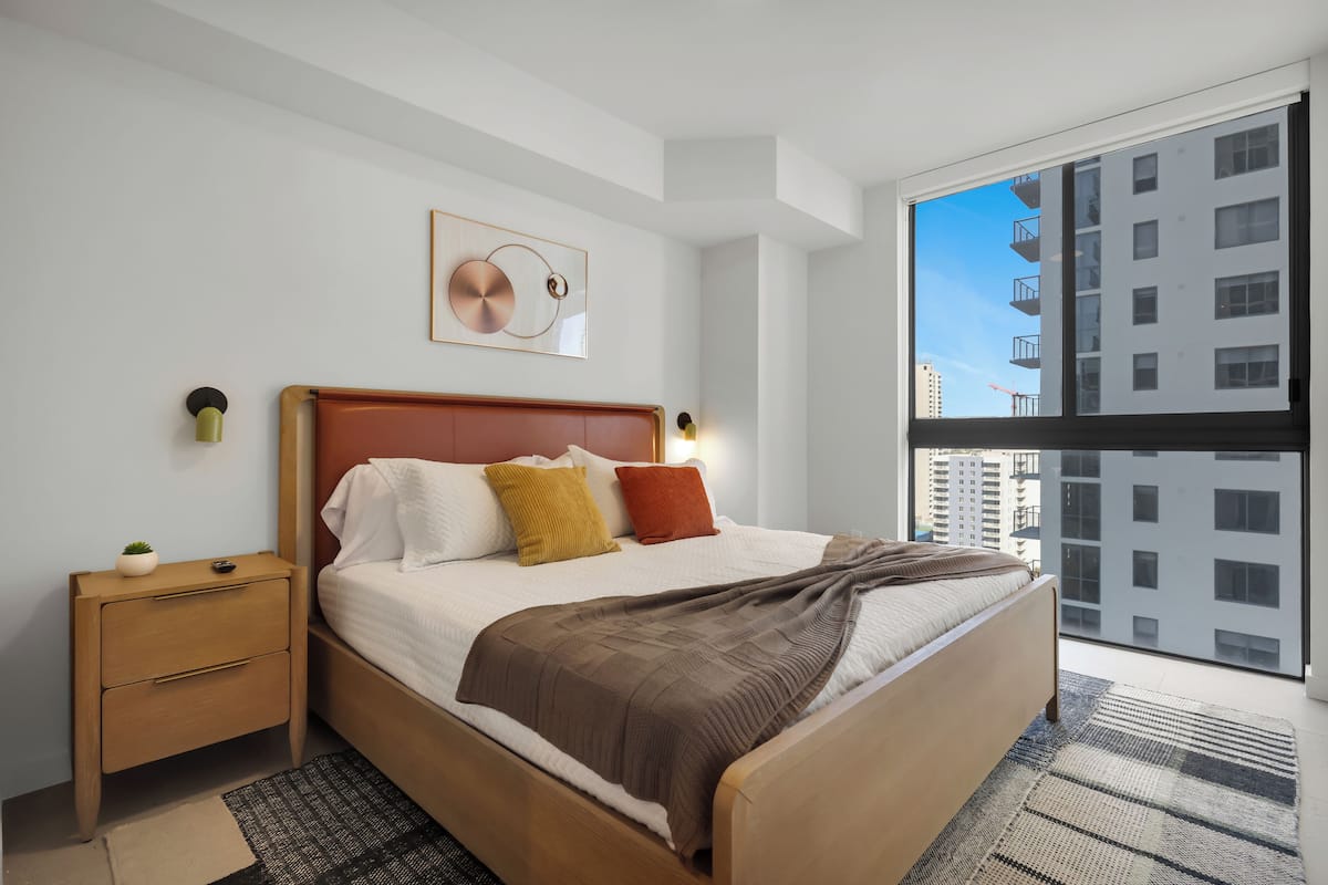 Una camera da letto moderna con un letto in legno, un comodino, una lampada a parete e un'ampia finestra con vista sulla città.