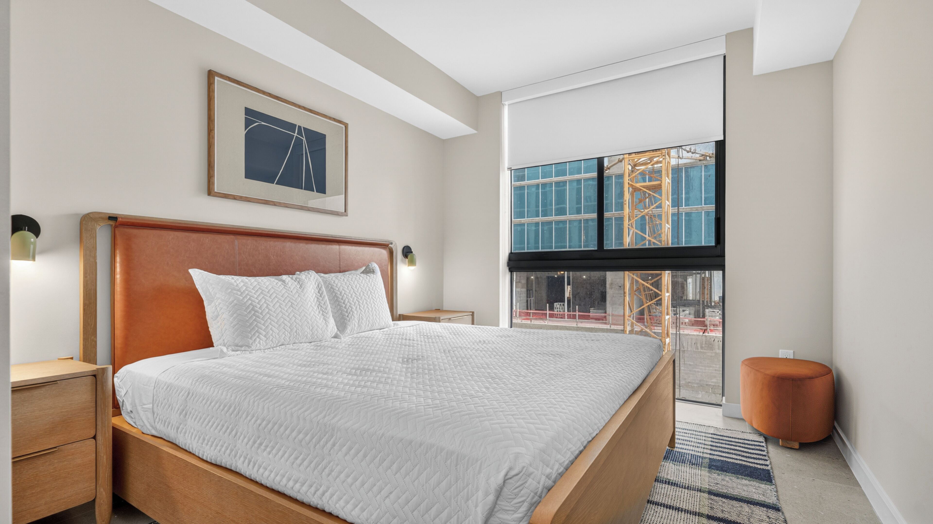 Ein Hotelzimmer mit einem großen Bett, einem gerahmten Bild an der Wand, einem Fenster mit Blick auf eine Baustelle und einem kleinen Nachttisch.