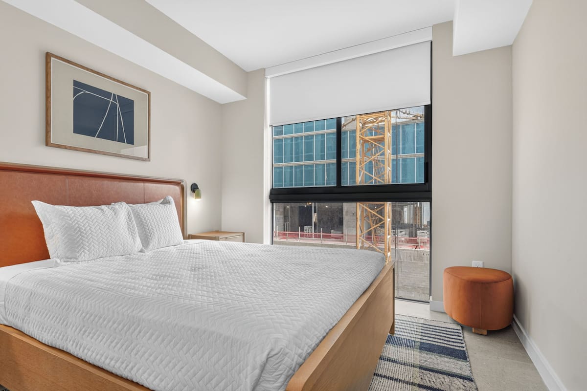Habitación de hotel con una cama grande, un cuadro enmarcado en la pared, una ventana con vista a una construcción y una mesita auxiliar pequeña.