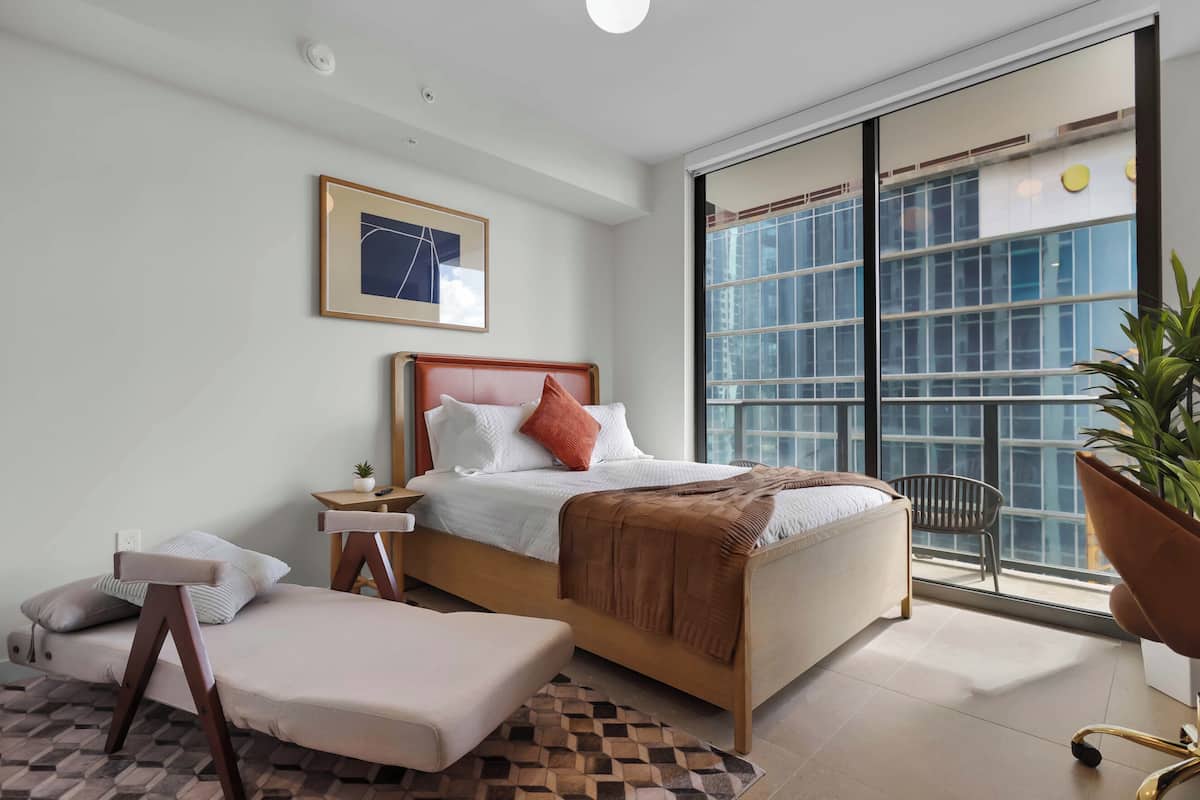 Un dormitorio moderno con cama, mesita de noche, silla y un ventanal con vistas a edificios.
