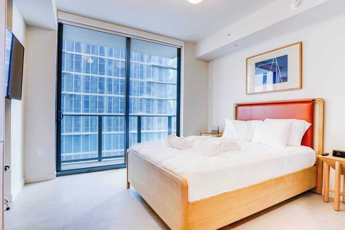 Un dormitorio con cama, televisor, mesita de noche, cabecera roja, ventana y un cuadro en la pared.