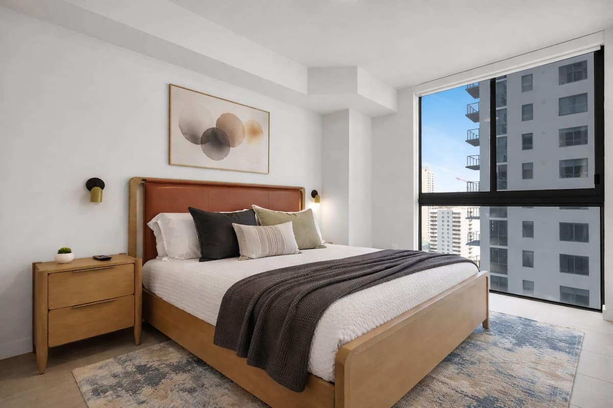 Un dormitorio moderno con un ventanal grande, una cama de madera, una mesita de noche y una lámpara fijada a la pared.