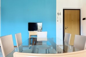 A room with a glass table, four chairs, a TV on a wooden cabinet, and a door with a 'Beach' sign. - P088 - Porto Recanati, new three-room apartment with balcony (Porto Recanati)