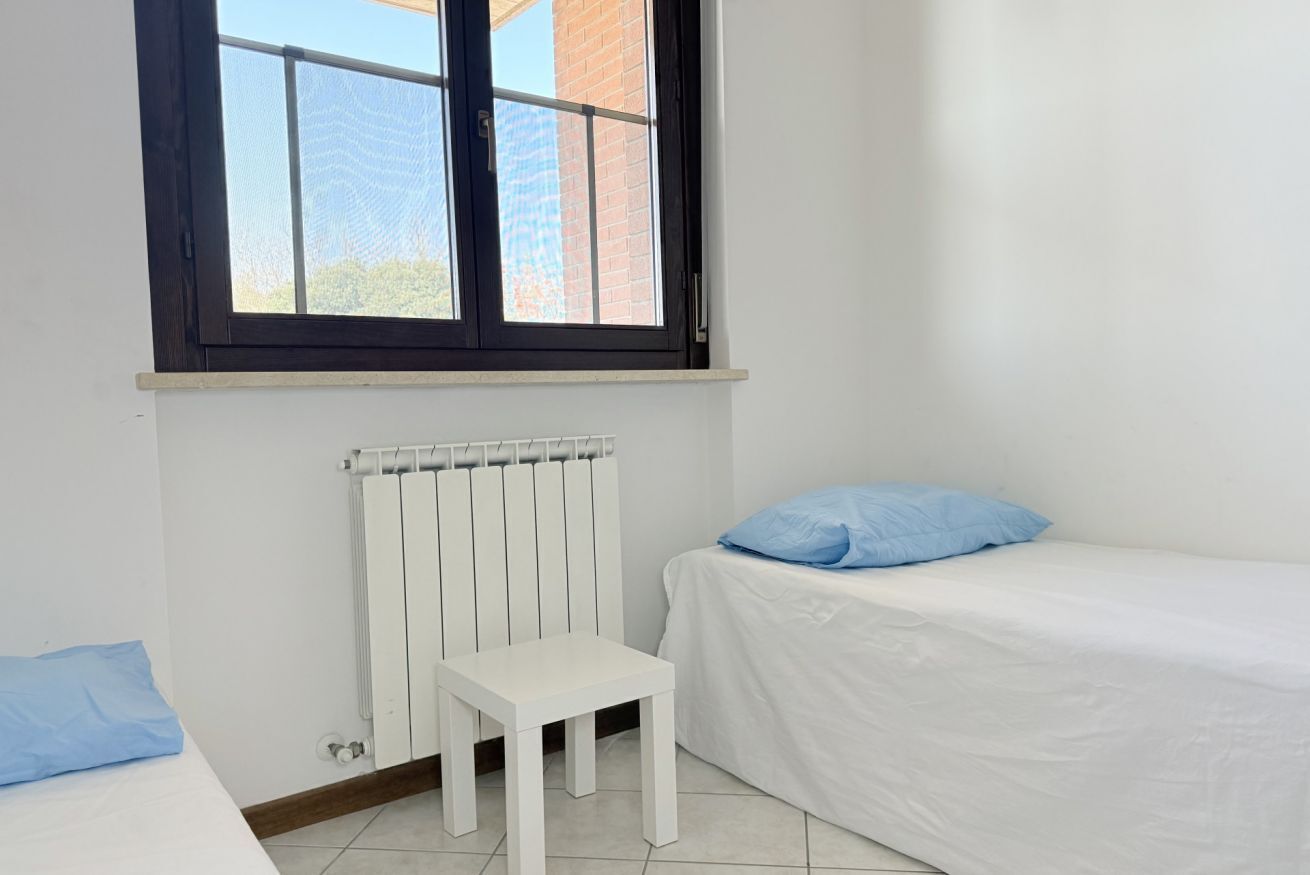 Una habitación con una cama, una mesita, un radiador y una ventana con vistas a un edificio.
