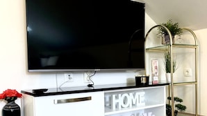 Ett modernt vardagsrum med en stor, platt-TV som är monterad på väggen, ett vitt konsolbord med lådor och en hylla i glas med växter.