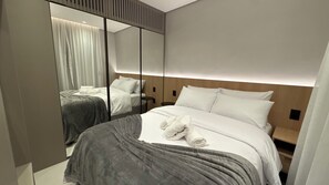 Ett modernt hotellrum med en stor spegel, en säng med vita sängkläder, ett grått överkast och en sänggavel i trä.