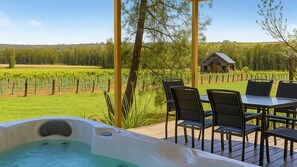 A hot tub on a wooden deck with a table and chairs, overlooking a vineyard and a rustic building. - La Capella Estate | On Shiraz Vineyard | Stunning Views (Pokolbin)