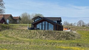 Ett modernt hus med stora fönster, ett sluttande tak och en grönskande kulle i förgrunden.
