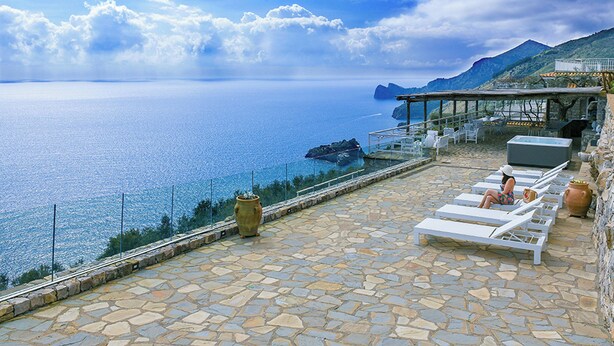 Eine Stegterrasse mit Liegestühlen mit Blick auf das Meer, eine Glasbrüstung und eine gebirgige Landschaft.