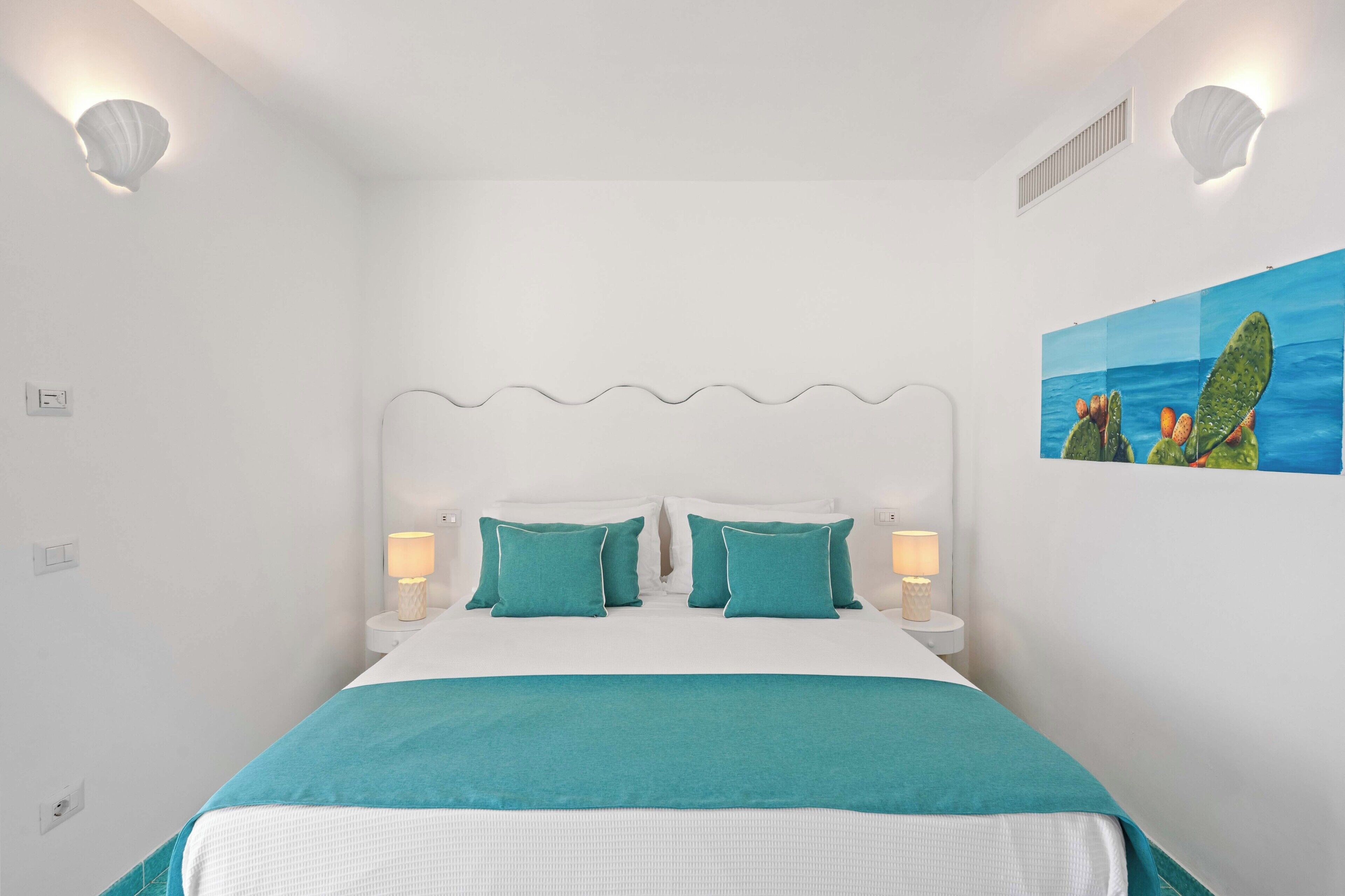 A bedroom with a bed, two bedside tables with lamps, a painting on the wall, and a ceiling vent.