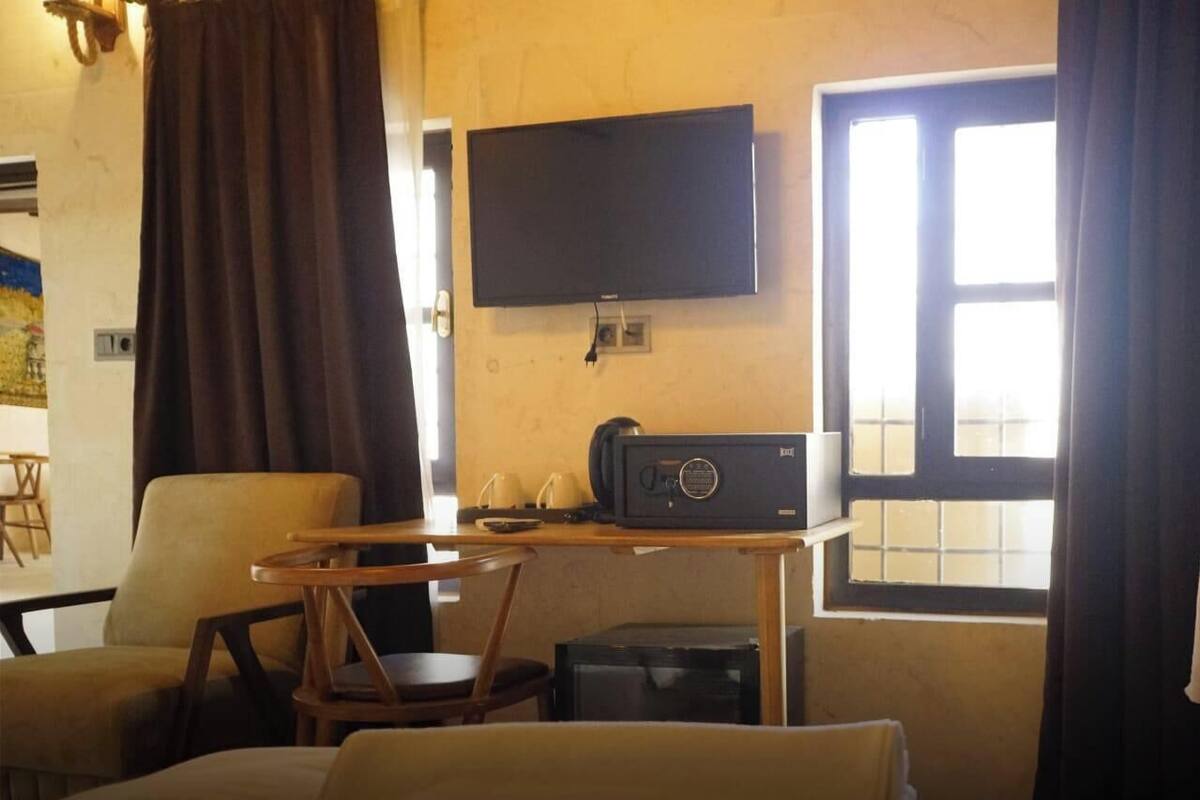 Quarto de hotel com televisão de tela plana, uma mesa pequena com um rádio, uma cadeira, uma cama e uma janela com cortinas.