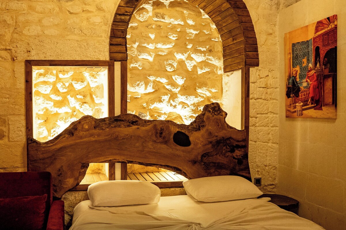 Una habitación con pared de piedra, cabecero de madera, cama con ropa blanca y una silla roja.