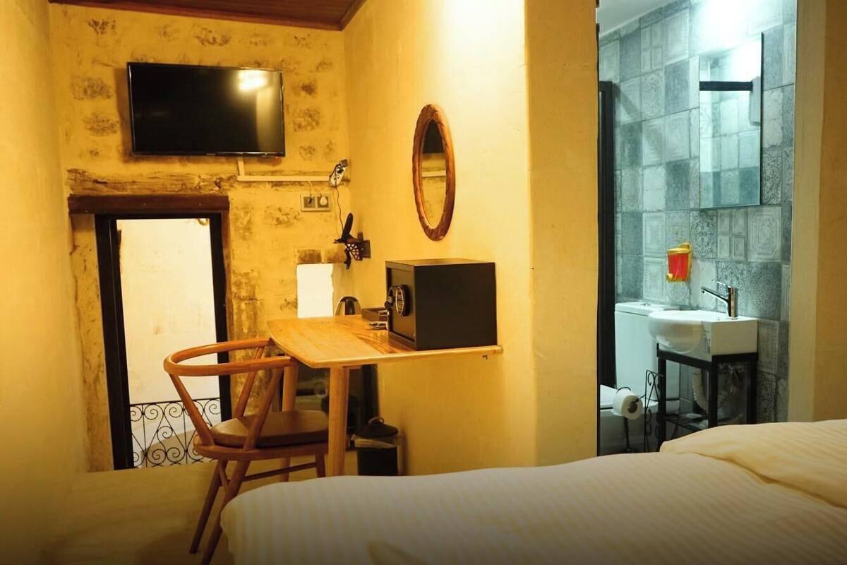 Quarto com cama, escrivaninha com cadeira, televisão, espelho e banheiro com pia e espelho.