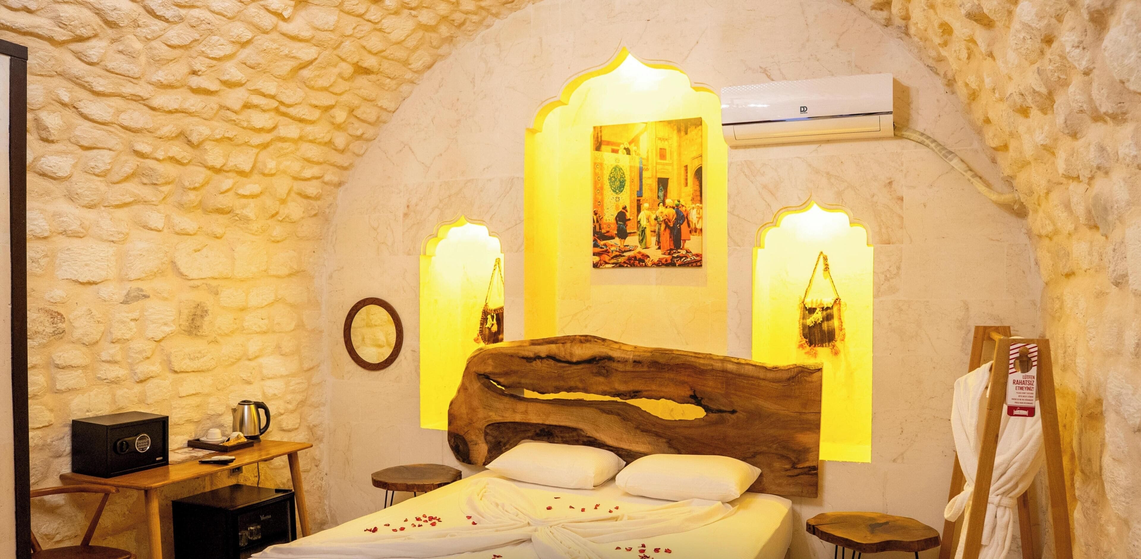 A room with a stone wall, a wooden bed, a nightstand, a table with a kettle, and a wall-mounted air conditioner.