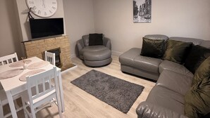 A living room with a fireplace, a sofa, a dining table with chairs, a TV mounted on the wall, and a clock. - Modern Comfort Home | Relax & Unwind in our 4 bed family friendly. (Oxford)