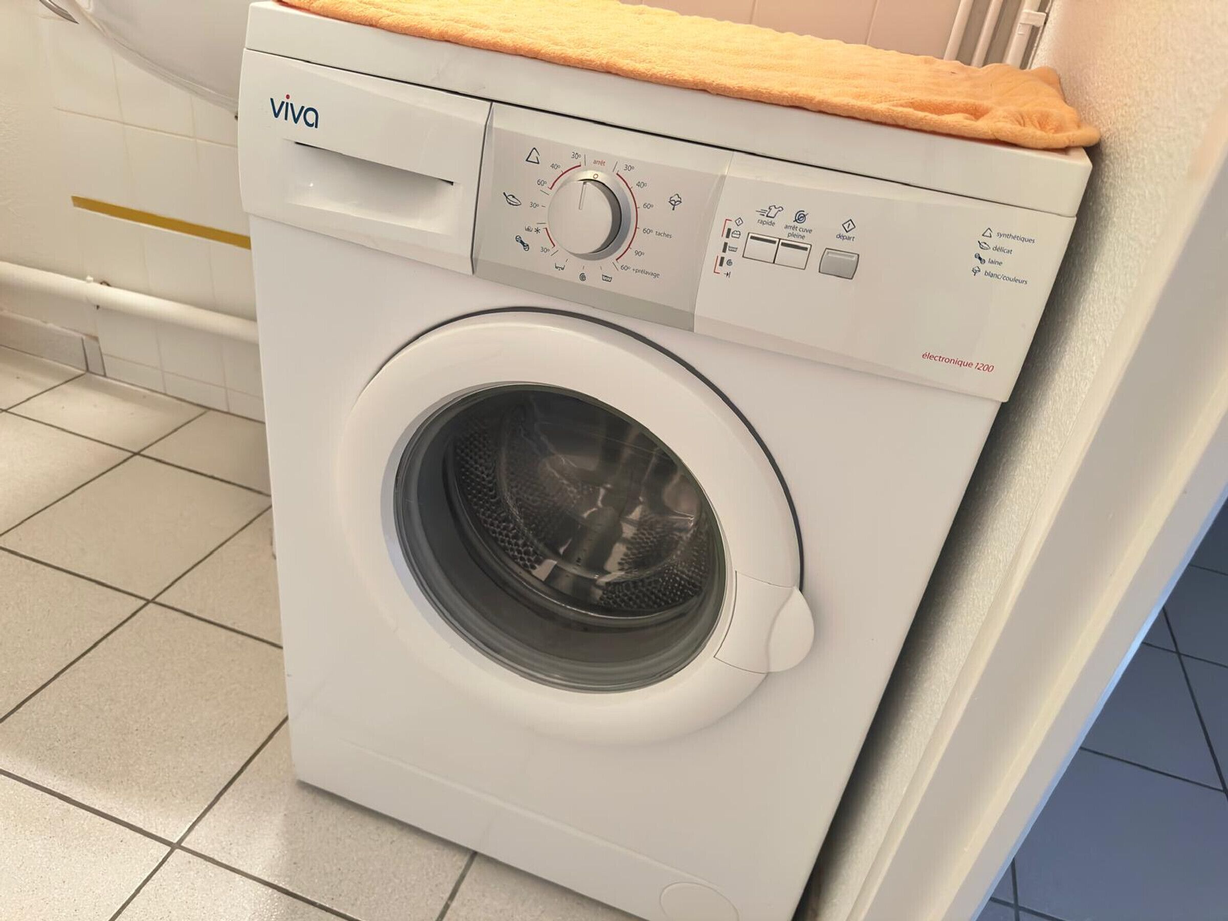 A front-loading white washing machine with a control panel, a wooden countertop, and a tiled floor.
