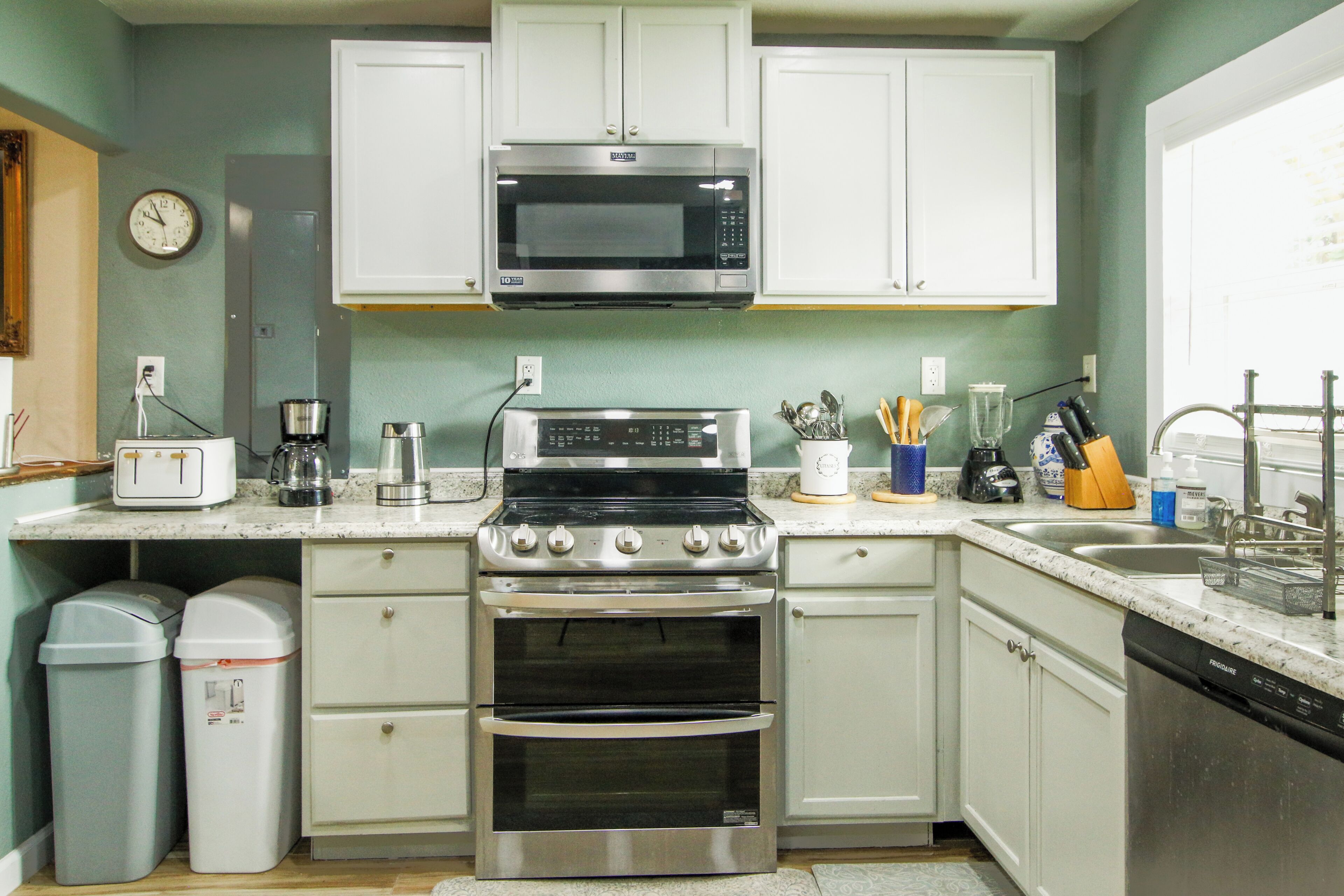 Una cocina con armarios blancos, una estufa de acero inoxidable, un microondas, un lavabo y varios electrodomésticos.