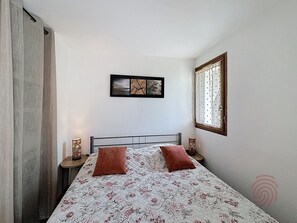 Een slaapkamer met een bed, twee kussens, een nachtkastje met een lamp, een lijst aan de muur en een raam met gordijnen.