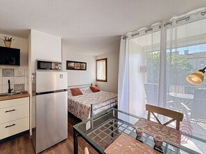 Een compacte kamer met een bed, een kleine kitchenette, een eettafel met stoelen en een groot raam met gordijnen.