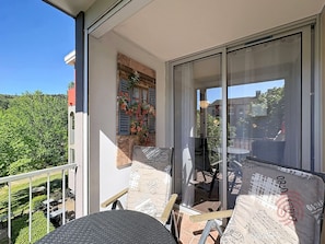 Een balkon met een tafel en stoelen, een raam met luiken, en uitzicht op groen.